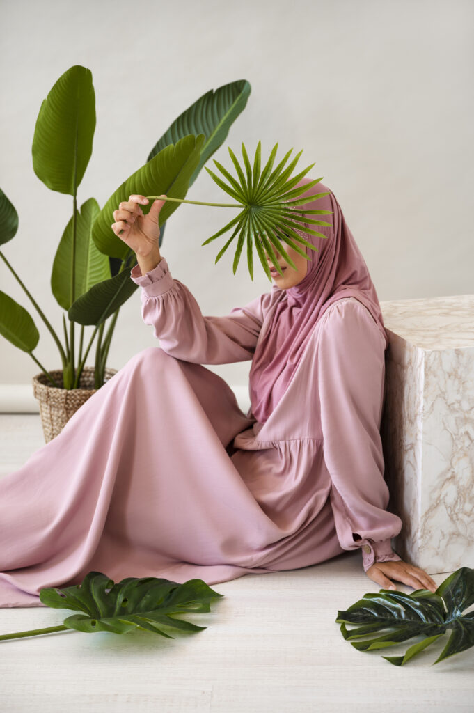 Femme portant une robe longue rose et un hijab assorti, posant dans un décor naturel avec des feuilles tropicales, tendances modest fashion